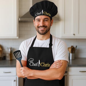 May include: A black chef's hat and apron set with the name "Chef Chris" printed in white and gold. The hat features a knife graphic. The apron has a chef's hat and knife graphic. A black spatula is held in the hand.