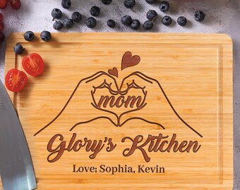 Könnte beinhalten: Ein rechteckiges Schneidebrett aus Bambus mit einer lasergravierten Abbildung von zwei Händen, die ein Herz um das Wort mom bilden. Darunter steht der Text Glory's Kitchen, gefolgt von den Namen Love; Sophia, Kevin in dunkelbrauner Schrift.