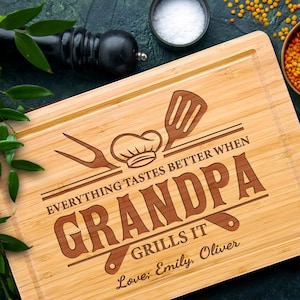 May include: A rectangular bamboo cutting board with a juice groove. The board features engraved text that reads "EVERYTHING TASTES BETTER WHEN GRANDPA GRILLS IT" with a chef's hat, spatula, and fork design. The names "Emily, Oliver" are also engraved.