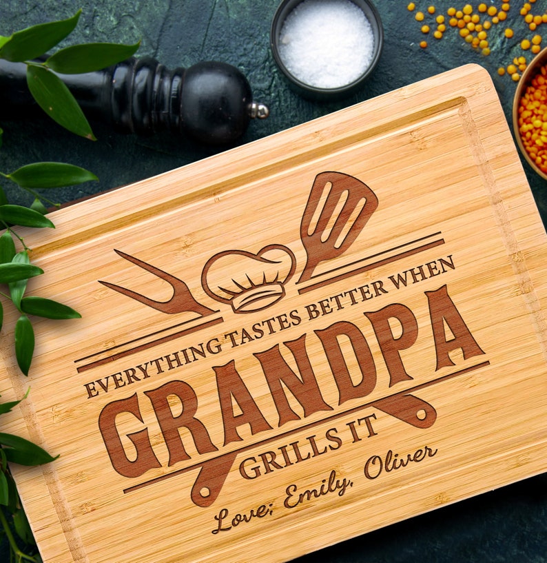 May include: A rectangular bamboo cutting board with a juice groove. The board features engraved text that reads "EVERYTHING TASTES BETTER WHEN GRANDPA GRILLS IT" with a chef's hat, spatula, and fork design. The names "Emily, Oliver" are also engraved.