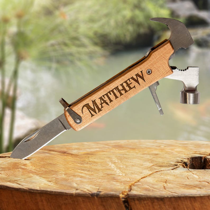 K&ouml;nnte beinhalten: Ein Multitool mit Holzgriff, mit Hammer, Messer und anderen Werkzeugen. Der Name "MATTHEW" ist auf dem Griff eingraviert. Das Werkzeug liegt auf einer Holzoberfl&auml;che, mit einem verschwommenen Hintergrund aus Gr&uuml;n und Wasser.