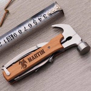 May include: A multi-tool hammer with a wooden handle, featuring a knife, pliers, and other tools. The handle is engraved with the name "MARTIN" and a dragon emblem. A metal tape measure and screws are also visible.