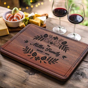 May include: A rectangular wooden cutting board with a dark brown finish. The board is engraved with the words "Miller Family Easter 2026" and decorative Easter-themed elements. Two glasses of red wine are in the background.