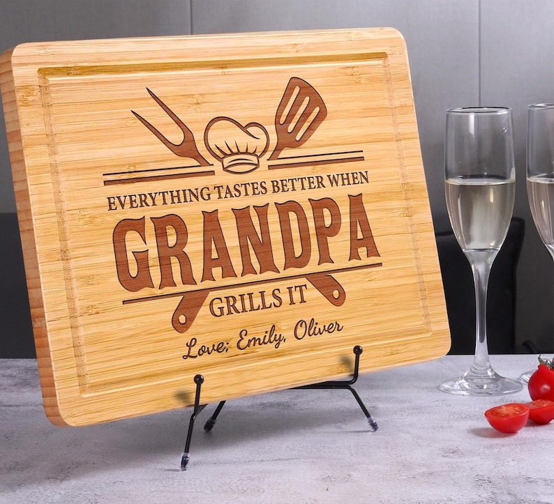 May include: A rectangular bamboo cutting board with a carved design. The text reads "EVERYTHING TASTES BETTER WHEN GRANDPA GRILLS IT" with a chef's hat, fork, and spatula graphic. Includes "Love; Emily, Oliver".