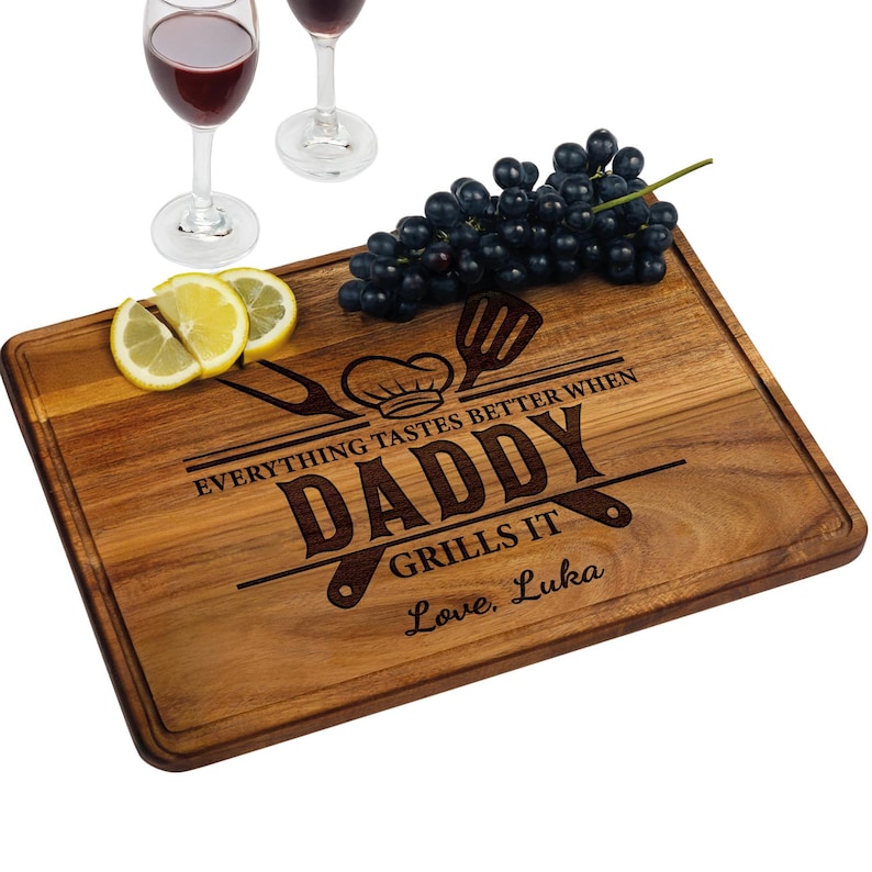 May include: A rectangular wooden cutting board with a juice groove, engraved with the words "DADDY GRILLS IT" and a chef's hat and utensils. Grapes and lemon slices are on the board, with two wine glasses in the background.