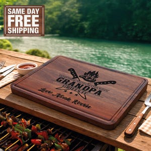 May include: A rectangular wooden cutting board with a dark brown finish. The board is engraved with the words "GRANDPA GRILLS IT" and "Love Noah, Kevin". The design includes images of knives and a grill. The board is on a wooden table.