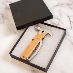 K&ouml;nnte beinhalten: Ein Multi-Tool-Hammer mit Holzgriff und Edelstahlkomponenten, pr&auml;sentiert in einer schwarzen Geschenkbox mit wei&szlig;em Innenraum. Der Hammer verf&uuml;gt &uuml;ber eine Klaue, ein Messer und andere Werkzeuge.