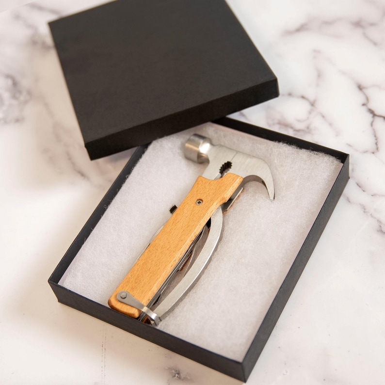 K&ouml;nnte beinhalten: Ein Multi-Tool-Hammer mit Holzgriff und Edelstahlkomponenten, pr&auml;sentiert in einer schwarzen Geschenkbox mit wei&szlig;em Innenraum. Der Hammer verf&uuml;gt &uuml;ber eine Klaue, ein Messer und andere Werkzeuge.