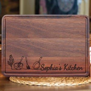 May include: A dark brown wooden cutting board with a design of a bowl, whisk, spatula, and pan engraved on the bottom. The text "Sophia's Kitchen" is engraved on the bottom of the board.