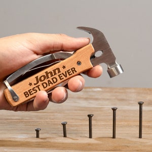 May include: A wooden multi-tool with a hammer head, a knife blade, and a screwdriver. The tool is engraved with the text "*John* BEST DAD EVER". The tool is being held by a hand and there are five nails on a wooden surface.