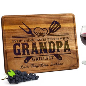 May include: Wooden cutting board with the text "Everything tastes better when Grandpa grills it." Includes a chef's hat, spatula, and fork design. Features the names "Love, Troy, Lisa, Jackson." A bunch of grapes and a glass of red wine are also in the image.
