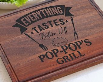 Könnte beinhalten: Ein rechteckiges, dunkelbraunes Holzschneidebrett mit einer Saftrille am Rand. Die Oberfläche zeigt eine schwarze Lasergravur mit dem Text EVERYTHING TASTES BETTER OFF POP-POP'S GRILL sowie Symbole einer Gabel, eines Pfannenwenders und einer Kochmütze.