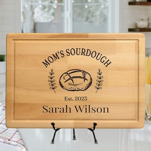 May include: A rectangular wooden cutting board with a decorative border. The board is engraved with "MOM'S SOURDOUGH", a bread illustration, "Est. 2023", and "Sarah Wilson". The board is displayed on a black metal stand.