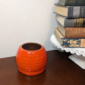 May include: A small, orange ceramic planter with a textured, woven design. The planter is empty and sitting on a wooden surface.