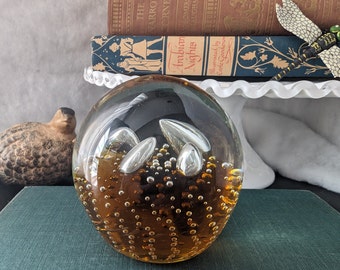 May include: A clear glass paperweight with a brown interior and silver bubbles. The paperweight has a textured surface and is filled with small, round bubbles.