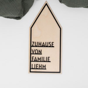 May include: A wooden house-shaped sign with the words "ZUHAUSE VON FAMILIE LIEHM" written in black lettering.