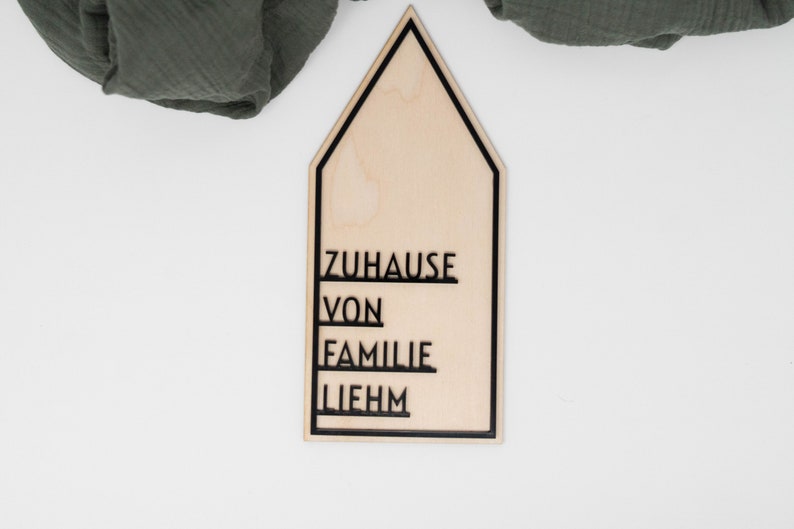 May include: A wooden house-shaped sign with the words "ZUHAUSE VON FAMILIE LIEHM" written in black lettering.