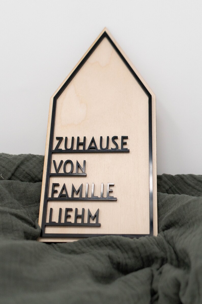 May include: A wooden house-shaped sign with black lettering that reads "Zuhause von Familie Liebhm".