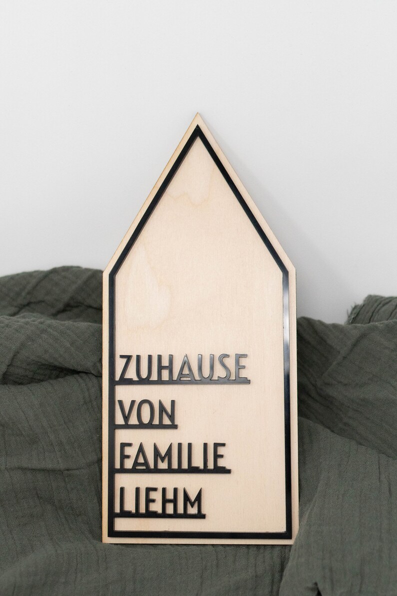 May include: A wooden house-shaped sign with black lettering that reads "ZUHAUSE VON FAMILIE LIEHM".