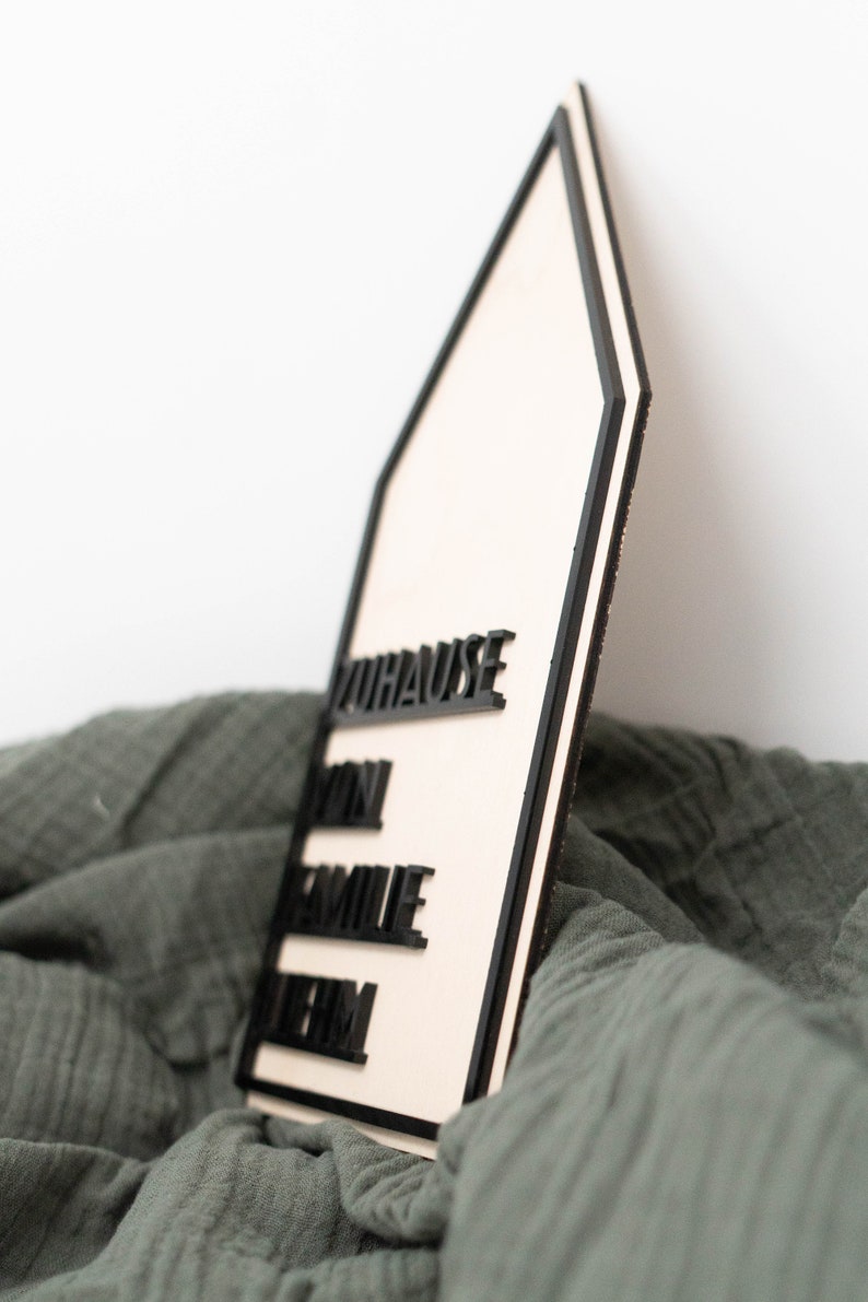 May include: A wooden house-shaped sign with black lettering that reads "Zuhause", "Dein", "Familie", and "Heim". The sign is sitting on a green fabric surface.