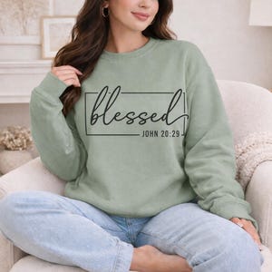 May include: Sage green sweatshirt with the word "blessed" in a black script font inside a black rectangle. Below the word "blessed" is the text "JOHN 20:29" in a smaller font. The sweatshirt is a crew neck style.
