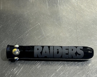 Puede incluir: Pipa para fumar de vidrio negro con la palabra "RAIDERS" grabada en el lateral. La pipa tiene detalles en vidrio azul y amarillo.