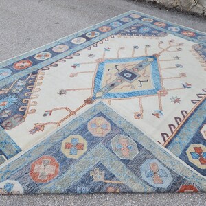 May include: A vintage rug with a cream background and a blue and orange geometric pattern. The rug has a diamond shape in the center with a smaller diamond inside. The rug is fringed on all sides.