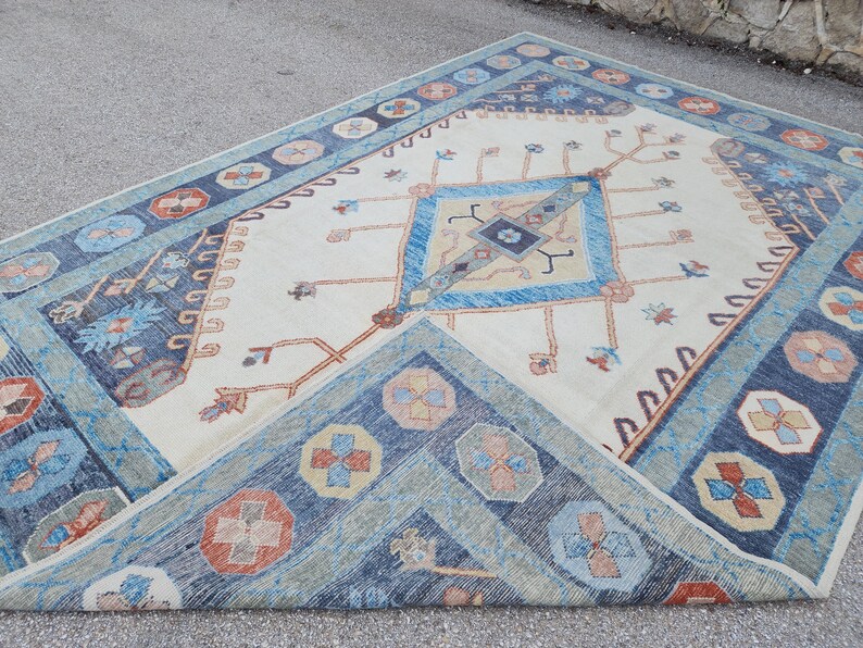 May include: A vintage rug with a cream background and a blue and orange geometric pattern. The rug has a diamond shape in the center with a smaller diamond inside. The rug is fringed on all sides.