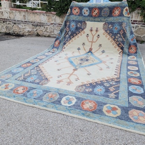 May include: A large, cream-colored rug with a blue and orange geometric pattern. The rug has a central diamond shape with a tree-like design and a border of repeating floral motifs.