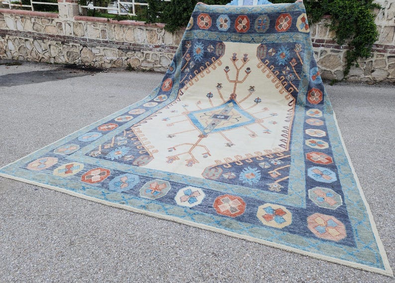 May include: A large, cream-colored rug with a blue and orange geometric pattern. The rug has a central diamond shape with a tree-like design and a border of repeating floral motifs.
