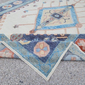 May include: A folded rug with a cream background and a blue, green, and orange geometric pattern. The rug has a fringed edge.