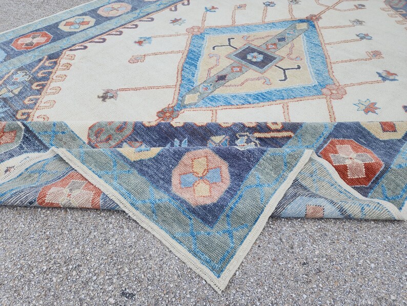 May include: A folded rug with a cream background and a blue, green, and orange geometric pattern. The rug has a fringed edge.