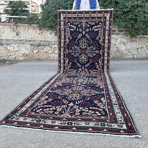 May include: A long, rectangular rug with an intricate floral pattern in navy blue, burgundy, and cream. The rug features a central medallion design and detailed borders. The rug is displayed outdoors, showcasing its length and design.