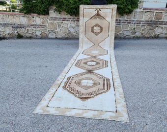 May include: A long, rectangular rug with a cream-colored background and geometric patterns in shades of brown. The rug features a central design with repeating diamond and hexagonal shapes, creating a symmetrical pattern. The edges are bordered with a darker brown trim.