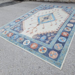 May include: A white rug with a blue and orange geometric pattern. The rug has a central diamond shape with a smaller diamond inside. The rug has a border of blue and orange flowers.