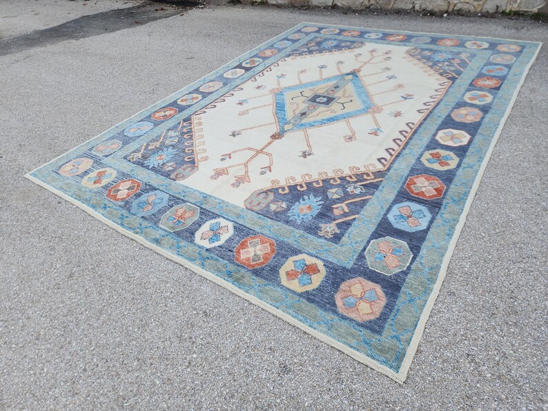 May include: A white rug with a blue and orange geometric pattern. The rug has a central diamond shape with a smaller diamond inside. The rug has a border of blue and orange flowers.