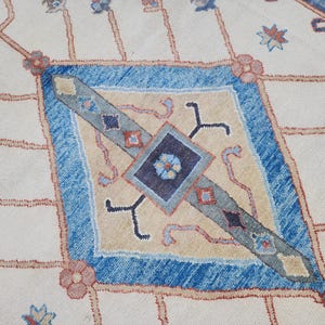 May include: A close-up of a cream-colored rug with a blue and beige diamond pattern. The diamond pattern features intricate details and a central square with a floral design.