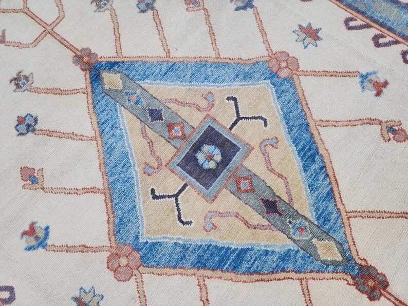 May include: A close-up of a cream-colored rug with a blue and beige diamond pattern. The diamond pattern features intricate details and a central square with a floral design.
