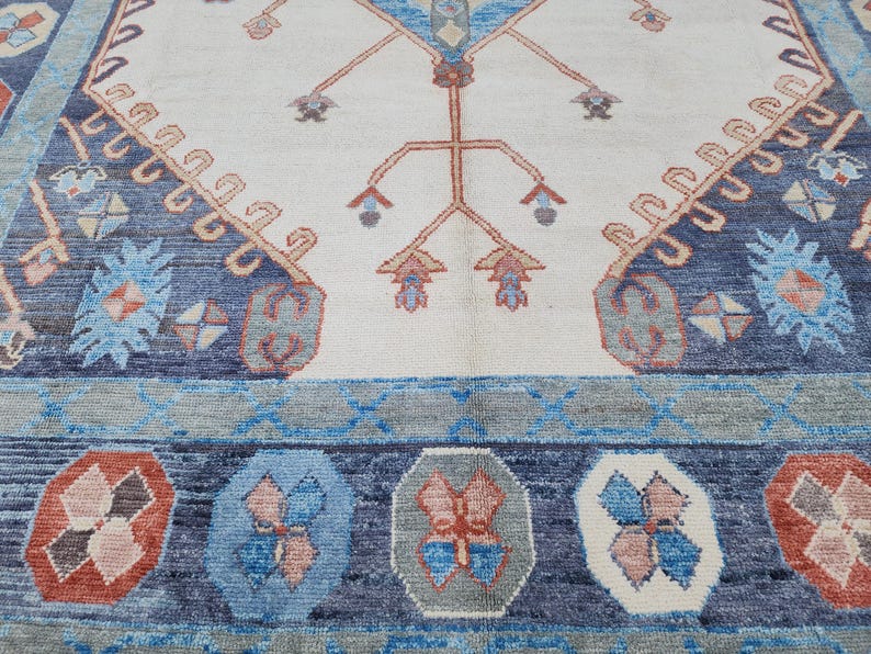 May include: Close-up of a rug with a blue and white border and a central design of a stylized tree with red and white accents.