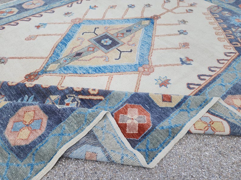 May include: A vintage rug with a cream background and a blue, red, and beige geometric pattern. The rug has a fringed edge and is folded in half.