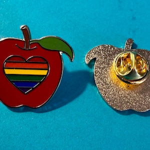 Può includere: Due spille a forma di mela in smalto. Una è rossa con una foglia verde e un design arcobaleno a forma di cuore. L'altra spilla è argentata con un retro dorato. Le spille sono su uno sfondo blu.