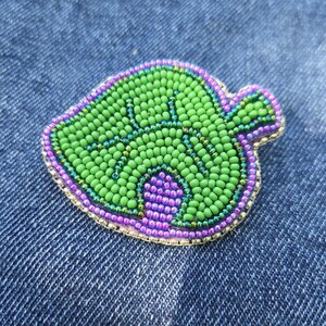 May include: A beaded leaf pin with a green centre, purple and gold trim, and a light green outline. The pin is made of small, round beads and is set against a blue denim background.