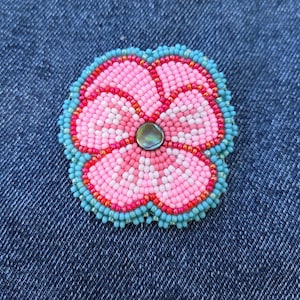 May include: A handmade beaded flower brooch featuring pink and white petals, a turquoise border, and an iridescent bead centre. The flower is set against a denim background.
