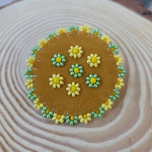 Peut inclure: Un écusson rond en daim marron orné de fleurs jaunes et vertes perlées. Les fleurs sont disposées en cercle, avec une bordure de petites perles jaunes et vertes. Cet article artisanal est idéal pour embellir des vêtements ou des accessoires.