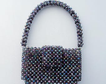 Puede incluir: Un pequeño bolso de mano de cuentas de color morado iridiscente con asa superior.