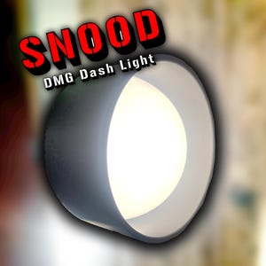May include: A circular, grey DMG Dash Light with a bright, white interior. The word "SNOOD" is displayed in red, outlined in black, above the light. The light is set against a blurred, neutral-toned background.