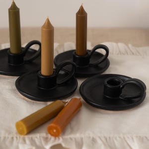 May include: Three black ceramic candle holders with a handle on each side. Each holder has a different colored candle, one green, one brown, and one tan. Two extra candles are on the white fabric surface.