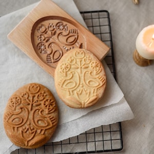 Puede incluir: Molde de galletas de madera con un diseño ovalado con pájaros y elementos florales, junto con dos galletas estampadas. Las galletas son de color marrón claro y el molde es de madera clara. También hay una vela encendida y utensilios para hornear.