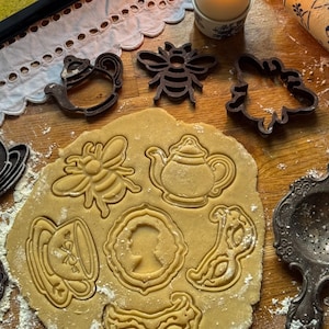 Puede incluir: Una imagen de naturaleza muerta de utensilios e ingredientes para hornear. Incluye una vela encendida, un rodillo y varios cortadores de galletas con forma de tetera, abeja y otros diseños decorativos. La masa de galletas ha sido cortada.