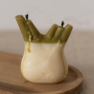 以下が含まれることがあります： 球根状の野菜の形をしたユニークなキャンドル。上部はオリーブグリーンで、アイボリー色のベースにワックスが垂れています。2本の消えた芯が見え、煙が立ち上っています。